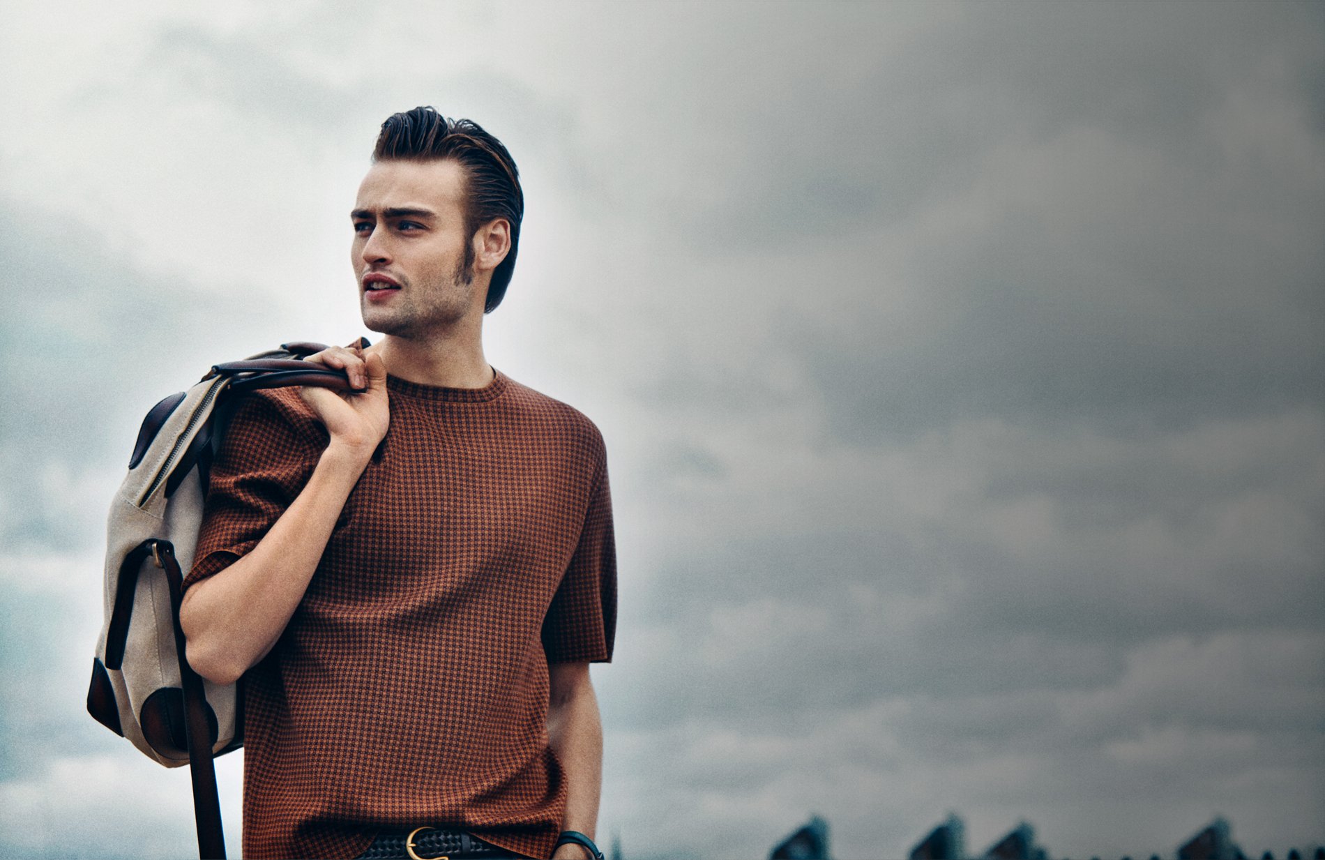 HD desktop wallpaper of English actor Douglas Booth wearing a brown sweater, holding a jacket over his shoulder against a cloudy sky backdrop.