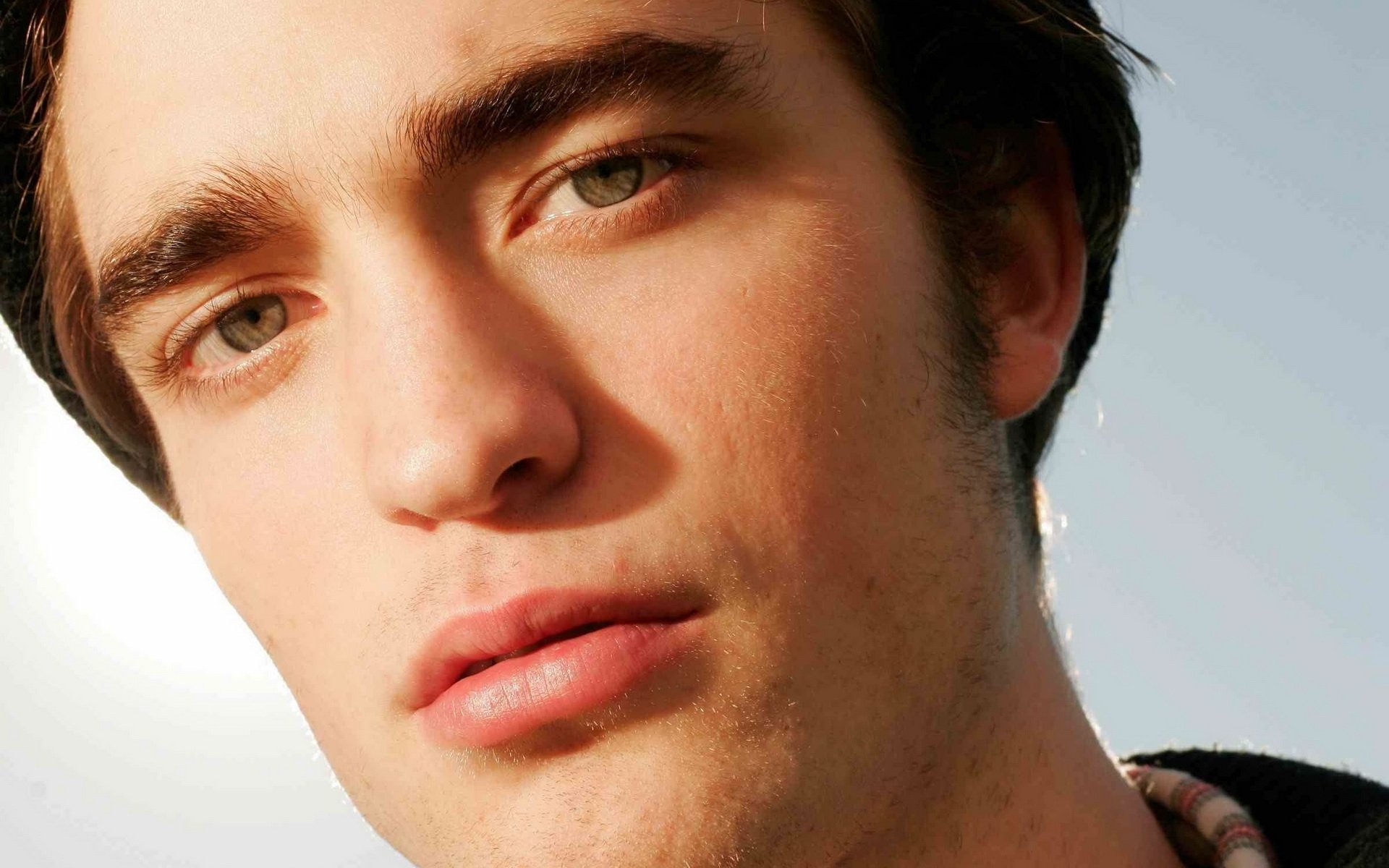 Close-up of an actor with striking blue eyes and a serious expression, set against a softly lit background. This high-definition wallpaper features Robert Pattinson.