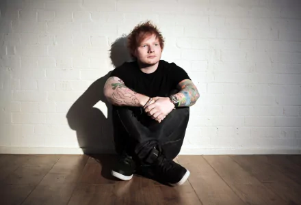 HD desktop wallpaper of English singer Ed Sheeran, showcasing his tattoos as he sits against a white brick wall, looking thoughtful.