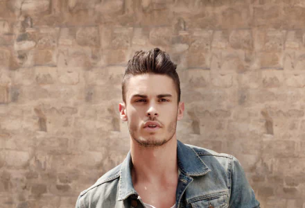 2K Quad HD desktop wallpaper of an unnamed French male model in a denim jacket posing against a sunlit stone wall.