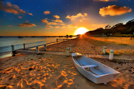 HD wallpaper showing a sunset over a sandy beach with a boat and a pier in the foreground and other boats in the distance. The sun is setting behind distant trees, casting warm hues over the sand and water.
