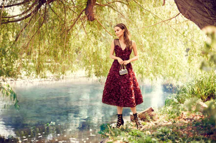 Alicia Vikander, a Swedish actress with brown eyes and brunette hair, poses gracefully by a serene waterway, wearing a stunning floral dress and holding a stylish bag.