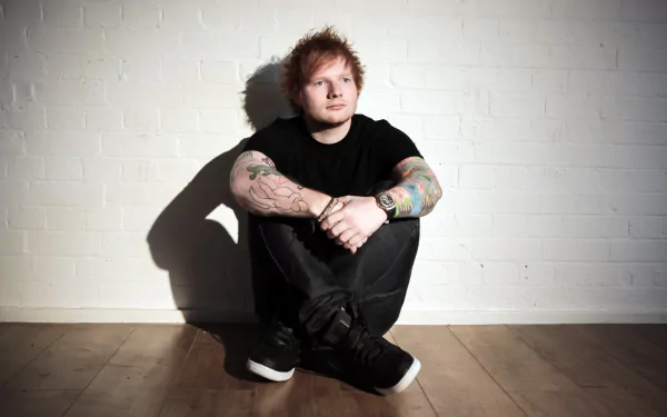 HD desktop wallpaper of English singer Ed Sheeran, showcasing his tattoos as he sits against a white brick wall, looking thoughtful.