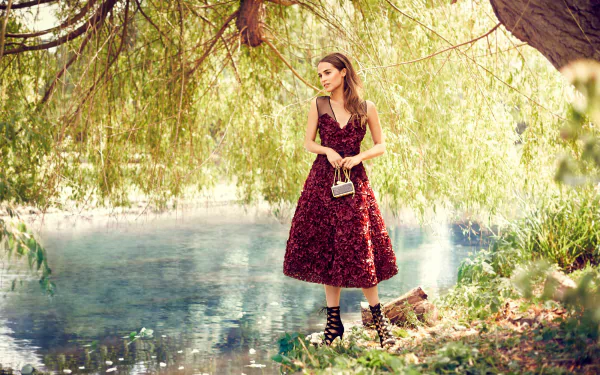 Alicia Vikander, a Swedish actress with brown eyes and brunette hair, poses gracefully by a serene waterway, wearing a stunning floral dress and holding a stylish bag.