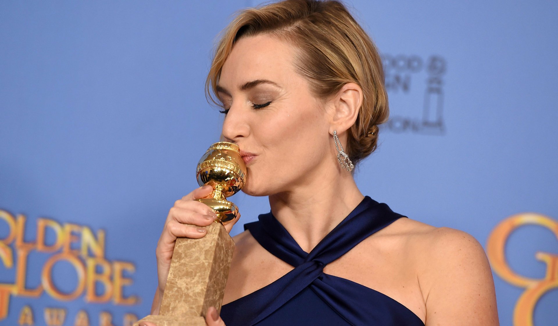 English actress Kate Winslet, with blonde hair and wearing earrings, kisses a Golden Globe award against a blue backdrop in this HD desktop wallpaper.
