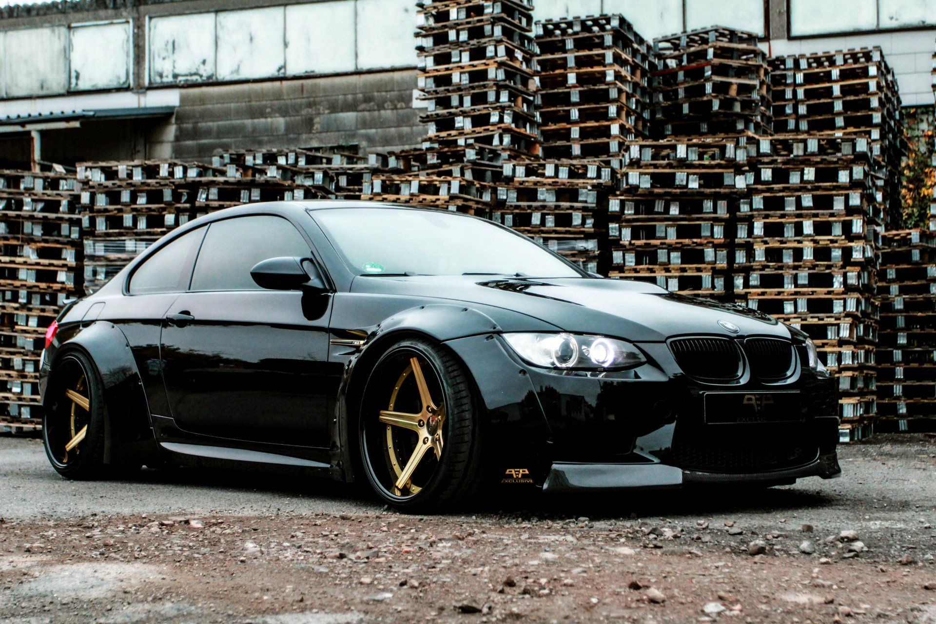 Sleek black BMW M3 Coupe with gold rims parked in front of stacked wooden pallets, captured in a high-definition desktop wallpaper.