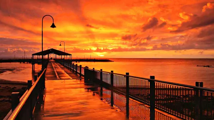 Sunrise over a man-made pier extending into the orange-hued ocean horizon in Queensland, Australia, with a vibrant sky reflecting on the wet walkway.