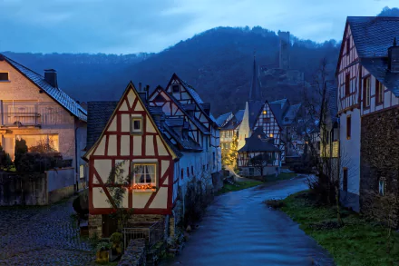 Dusk settles over a picturesque German village, showcasing charming architecture and cozy houses along a quiet winding road, with rolling hills in the background.