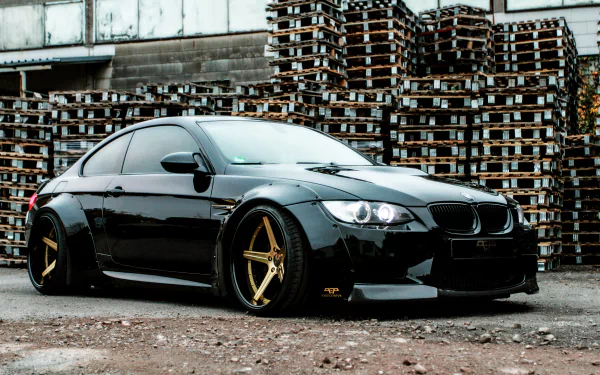 Sleek black BMW M3 Coupe with gold rims parked in front of stacked wooden pallets, captured in a high-definition desktop wallpaper.
