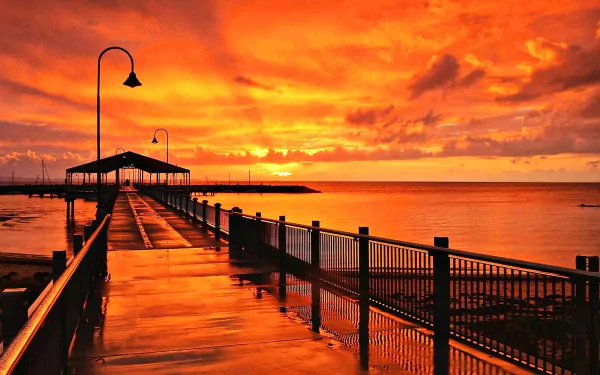 Sunrise over a man-made pier extending into the orange-hued ocean horizon in Queensland, Australia, with a vibrant sky reflecting on the wet walkway.