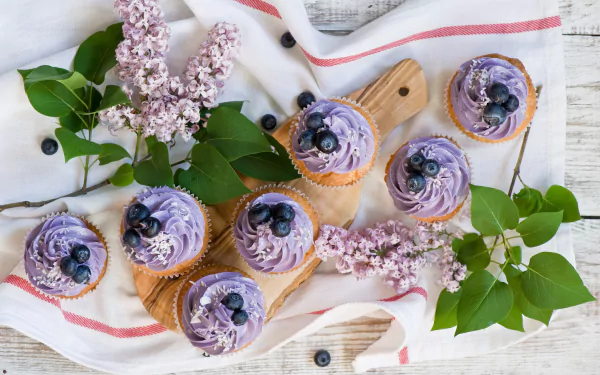 A 4K Ultra HD desktop wallpaper featuring lilac-colored blueberry cupcakes arranged on a wooden board with fresh green leaves and lilac flowers.