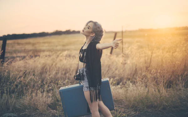 A woman with brown eyes poses outdoors in the sun, holding a suitcase and a camera, capturing a relaxed, contemplative mood in this 4K Ultra HD wallpaper.