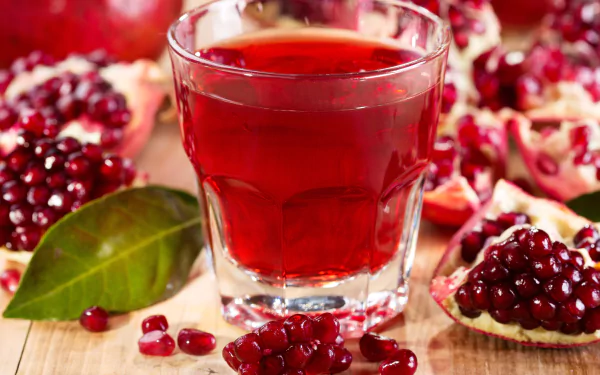 4K Ultra HD PC desktop wallpaper background: glass of ruby pomegranate juice with pomegranate halves and scattered seeds on a wooden board, food photography.