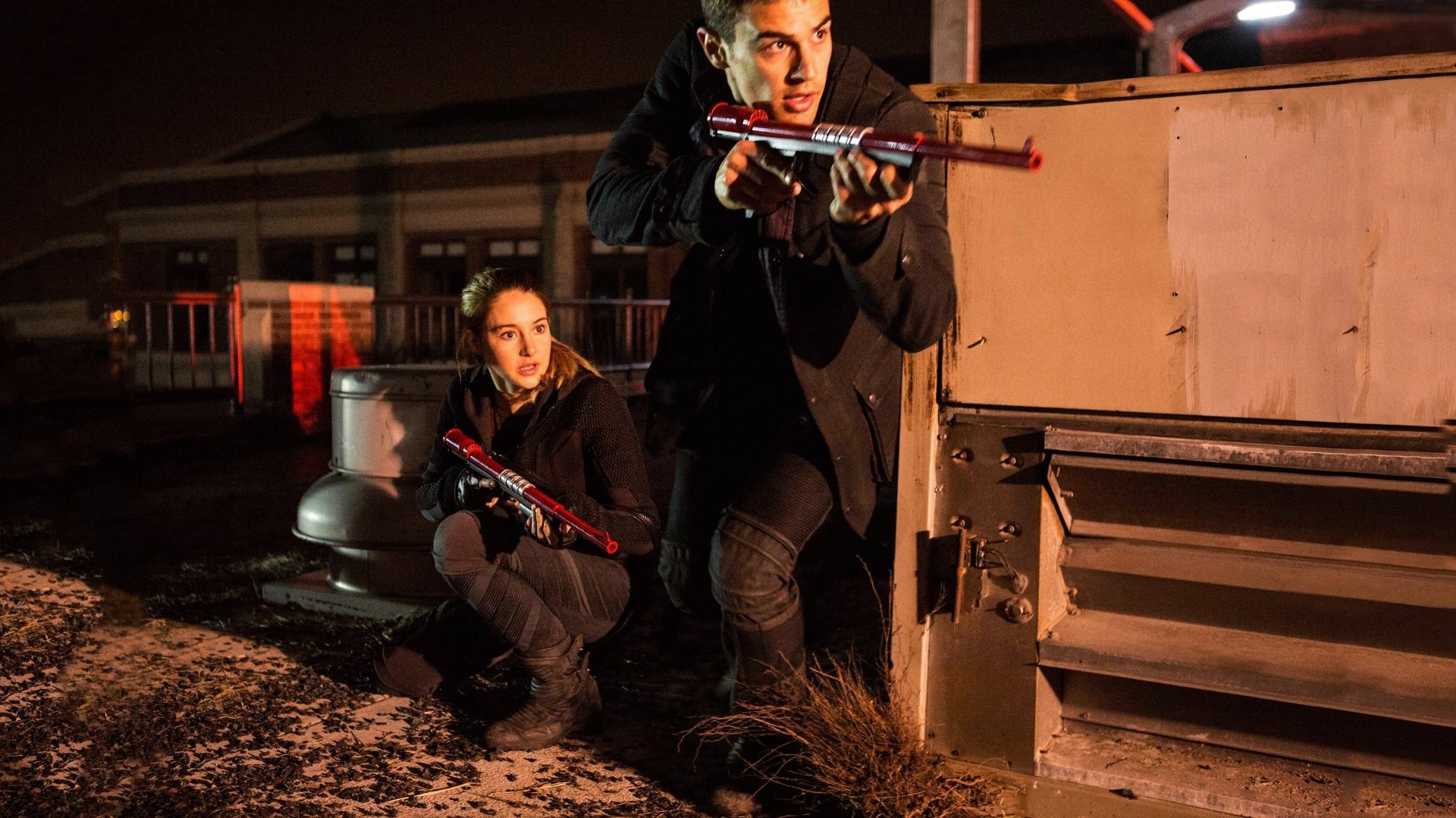 Two characters from the movie Divergent are depicted crouching on a rooftop at night, armed and ready for action, against a dramatic urban backdrop. HD desktop wallpaper.