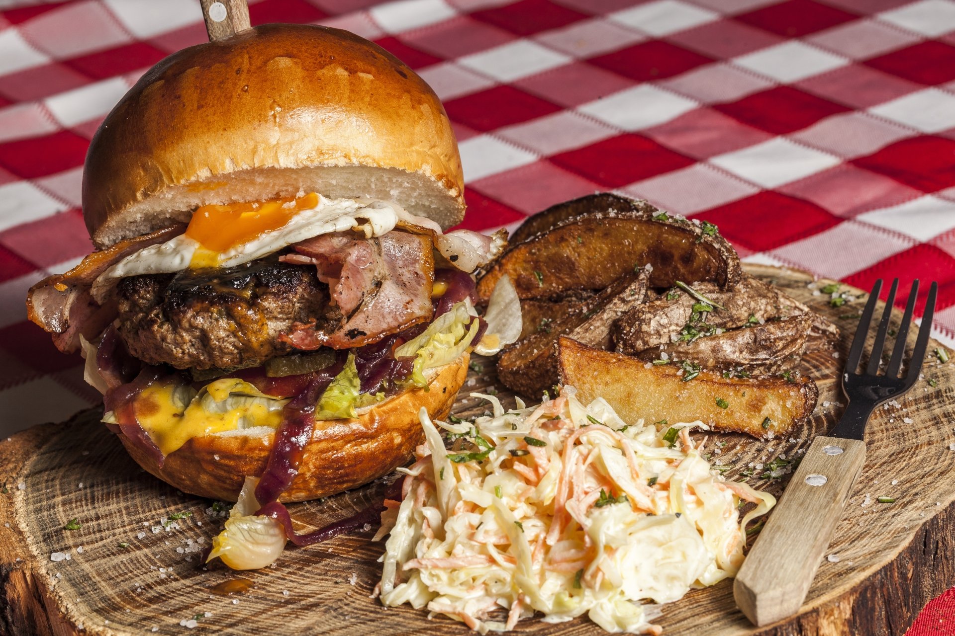 A 4K Ultra HD desktop wallpaper showing a hearty hamburger with egg and bacon, served with coleslaw and grilled mushrooms on a wooden board over a red checkered tablecloth.