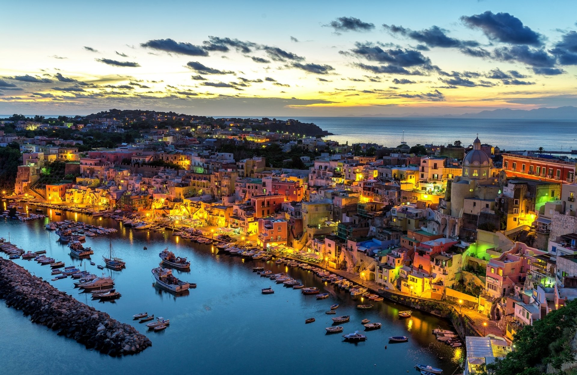 A vibrant dusk cityscape of Procida, Italy, featuring a bustling harbor with boats and illuminated town buildings along the waterfront.