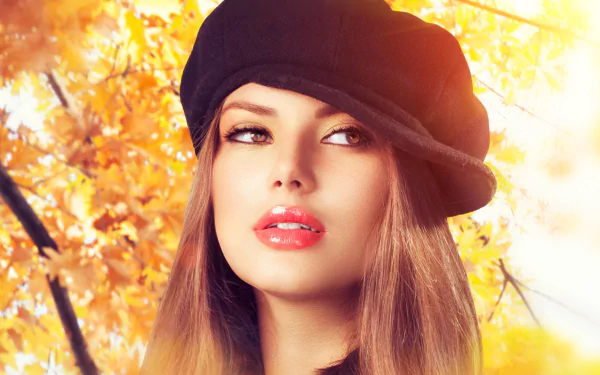 A woman with brown eyes and brunette hair wears a stylish hat against a backdrop of vibrant autumn leaves, illuminated by warm sunshine.