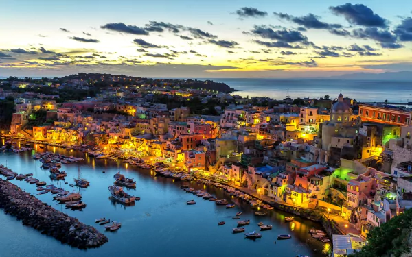A vibrant dusk cityscape of Procida, Italy, featuring a bustling harbor with boats and illuminated town buildings along the waterfront.
