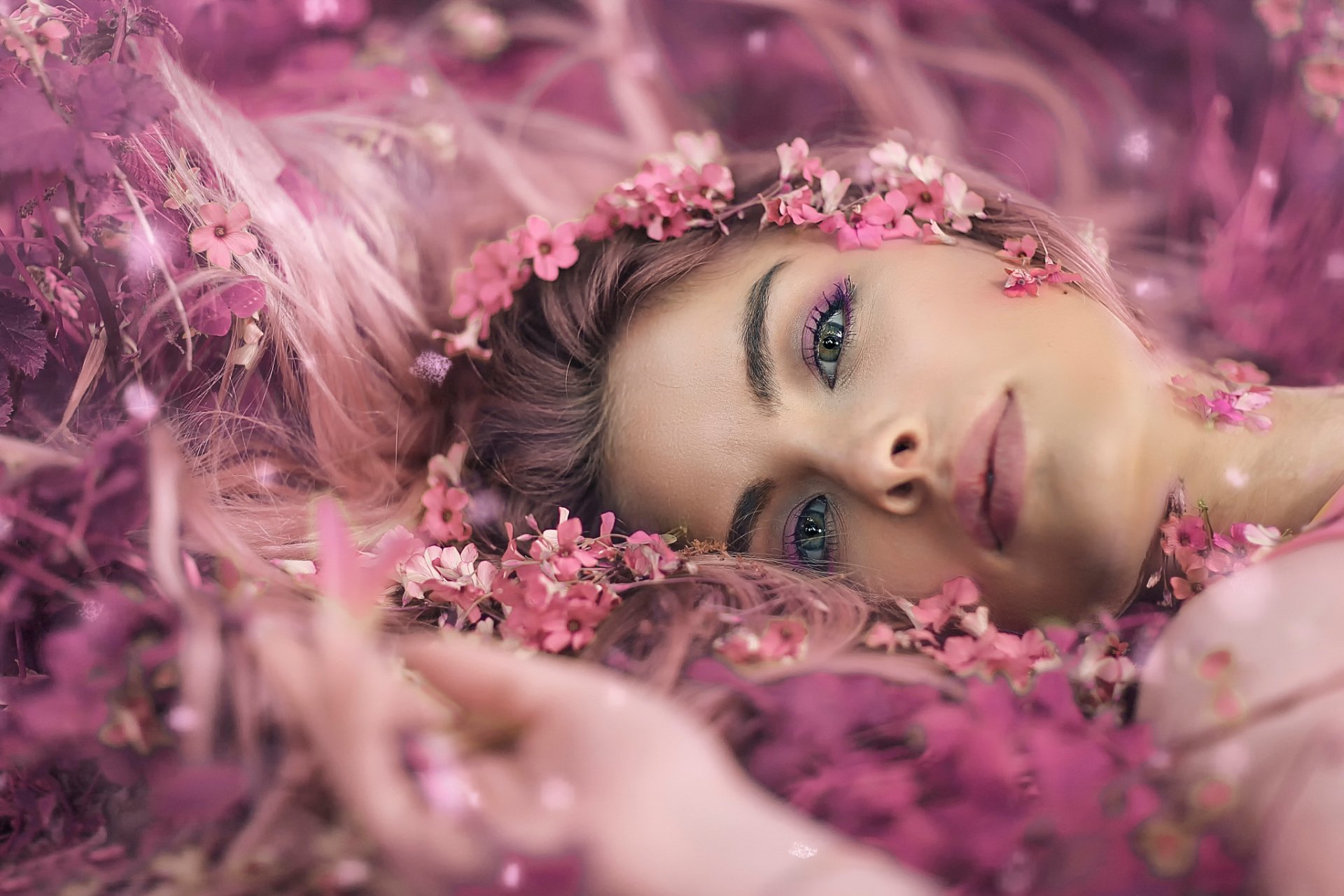 A woman with green eyes is lying down amidst pink flowers, creating a dreamy mood. This HD wallpaper captures her face surrounded by blossoms, making for an enchanting desktop background.