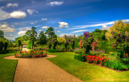 An HDR HD wallpaper featuring a vibrant spring garden in a park, showcasing colorful flowers and lush trees. A pathway winds through the well-maintained, man-made landscape under a bright blue sky.