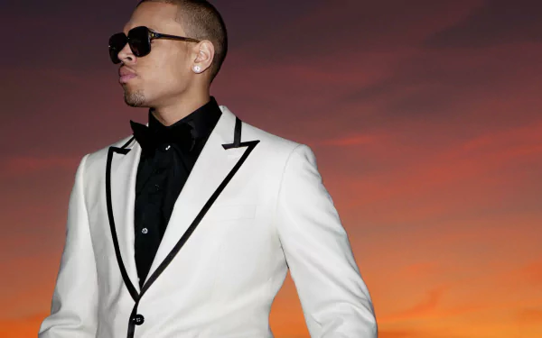 A stylish Chris Brown in a white suit stands against a vibrant sunset, capturing a dynamic moment in music. This HD wallpaper adds a touch of flair to any desktop background.
