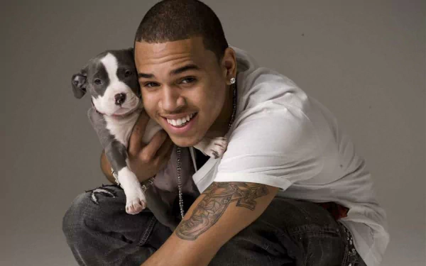 A high-definition desktop wallpaper featuring Chris Brown, smiling and posing with a small dog, showcasing his playful side against a neutral background.