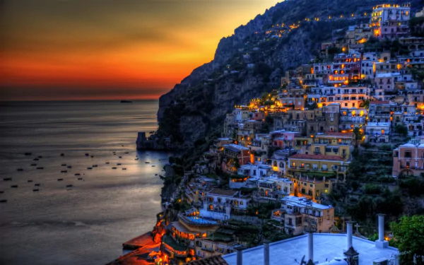 HDR night view of Positano, Italy, showcasing illuminated houses cascading down the coastal cliffs by the ocean, captured as a vibrant HD desktop wallpaper background.