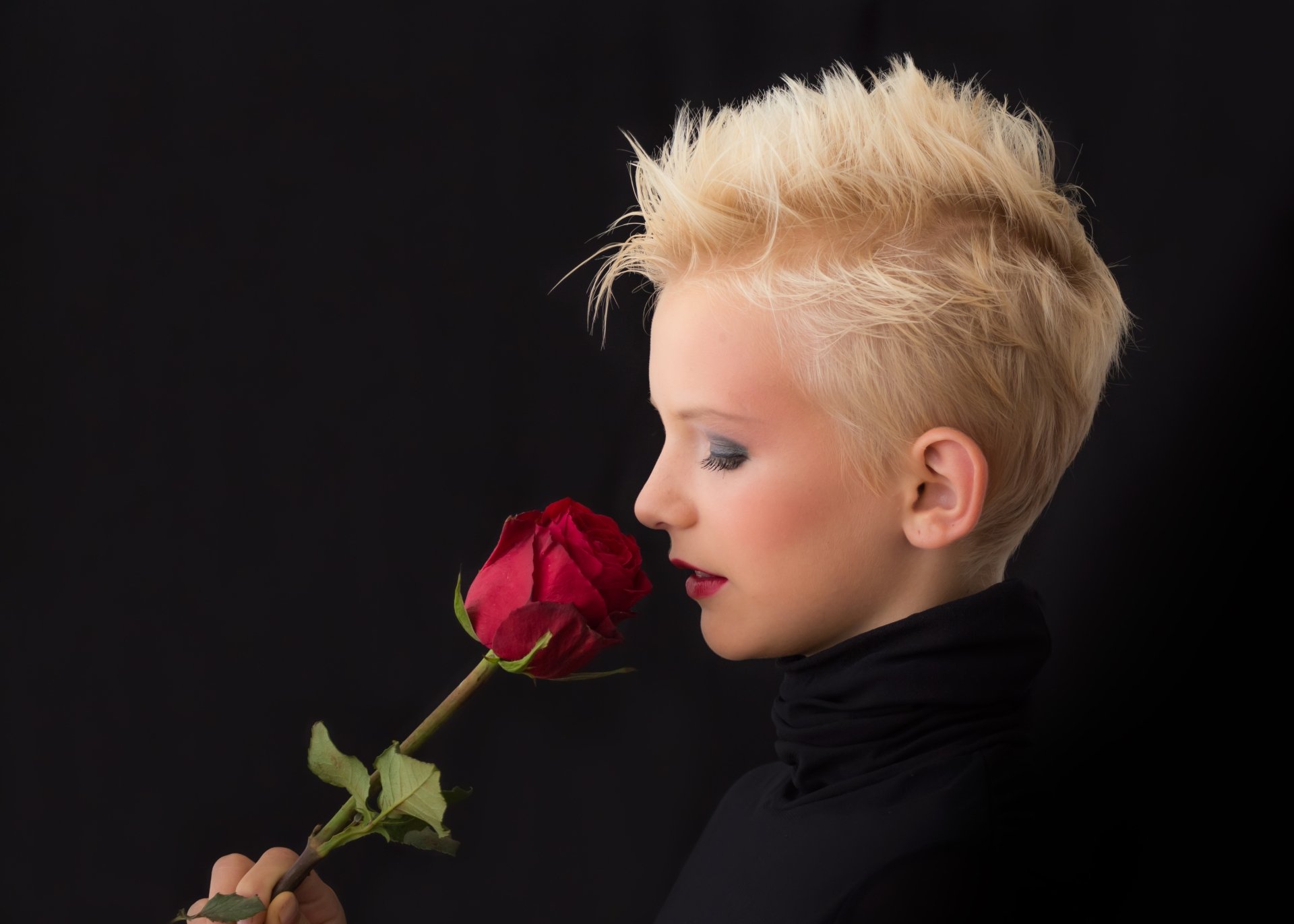 Blonde woman with bold makeup and red lipstick holding a red rose close to her face against a black background in 4K Ultra HD.