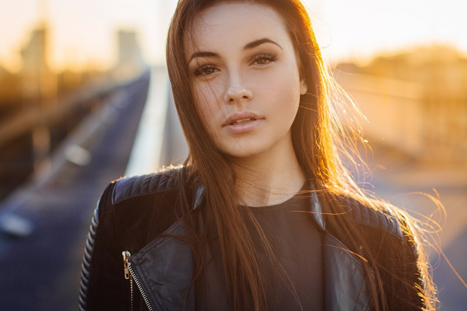 HD desktop wallpaper featuring a close-up portrait of a woman model with long hair, wearing a black leather jacket, bathed in warm golden sunlight.