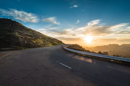 4K Ultra HD PC desktop wallpaper: winding man-made highway curves around a sunlit hill and mountain beneath a cloud-streaked sky with golden sunrise/sunset glow.