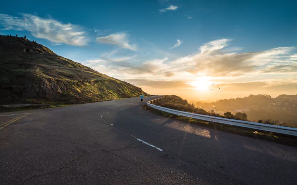 4K Ultra HD PC desktop wallpaper: winding man-made highway curves around a sunlit hill and mountain beneath a cloud-streaked sky with golden sunrise/sunset glow.