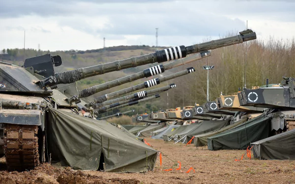 HD PC desktop wallpaper showing a row of Challenger 2 main battle tanks with raised guns at a muddy military encampment beneath a cloudy sky.