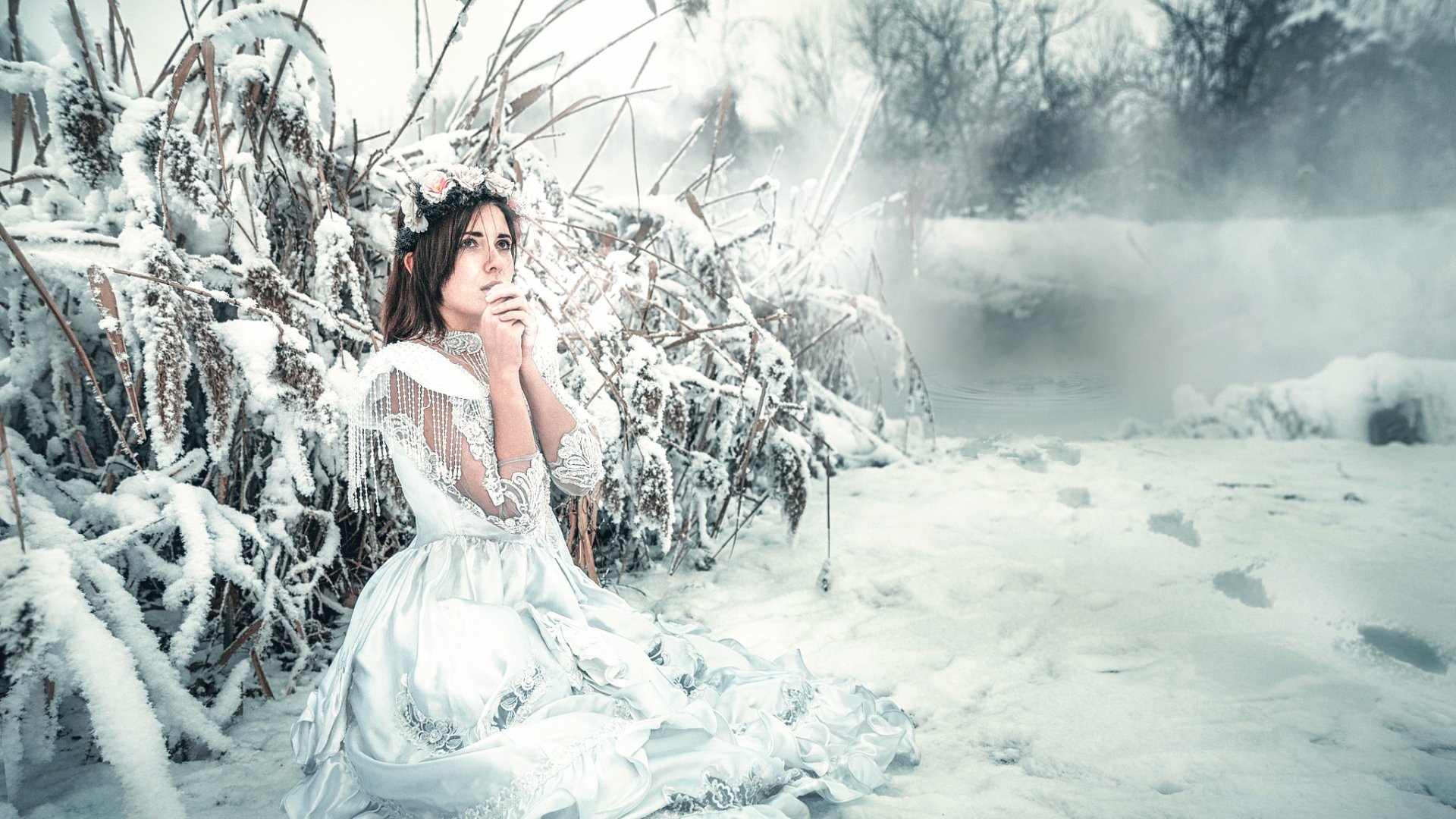 A brunette woman in a white dress and floral wreath sits thoughtfully in a snowy winter landscape, creating a serene and moody outdoor scene.