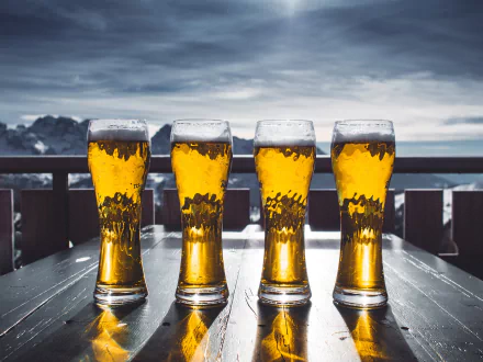 Four tall glasses of beer with frothy heads sit on a wooden table outdoors under a cloudy sky, captured in crisp 4K Ultra HD for a PC desktop background.