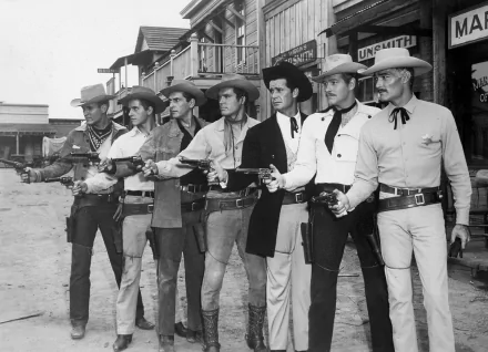 A black and white HD desktop wallpaper featuring a group of Western-clad actors holding guns, set in a rustic town backdrop. The image captures a classic cinematic moment.
