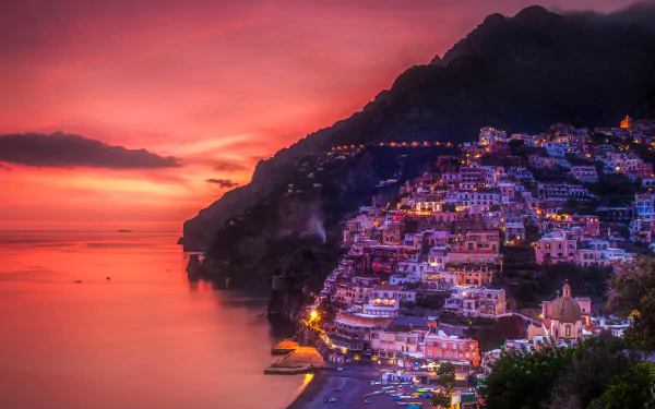 A stunning 4K Ultra HD view of Positano, Italy, showcasing illuminated houses along the coastal cliffs at sunset with vibrant, colorful sky and calm sea.