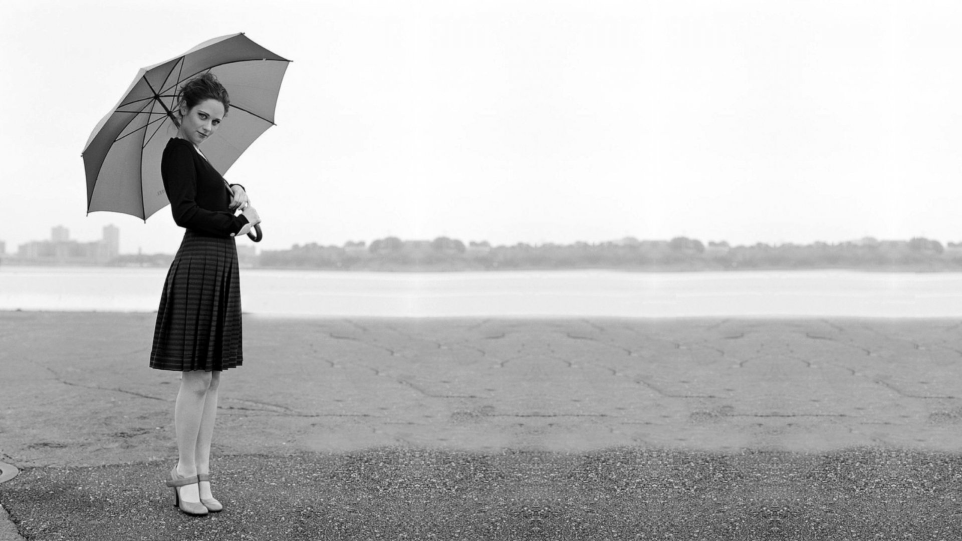 A black and white HD desktop wallpaper featuring Zooey Deschanel, elegantly holding an umbrella while standing by a calm shoreline.