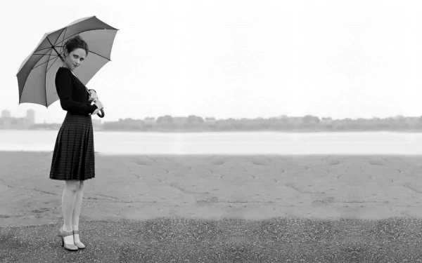 A black and white HD desktop wallpaper featuring Zooey Deschanel, elegantly holding an umbrella while standing by a calm shoreline.