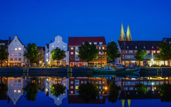 HD PC desktop wallpaper: night view of Lübeck, Germany — historic houses and twin church spires mirrored in calm water, a man-made town waterfront reflection under a deep blue sky.