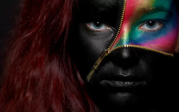 A woman with striking blue eyes and red hair, her face painted black with a leather-like texture, partially unzipped to reveal vibrant, colorful paint beneath.