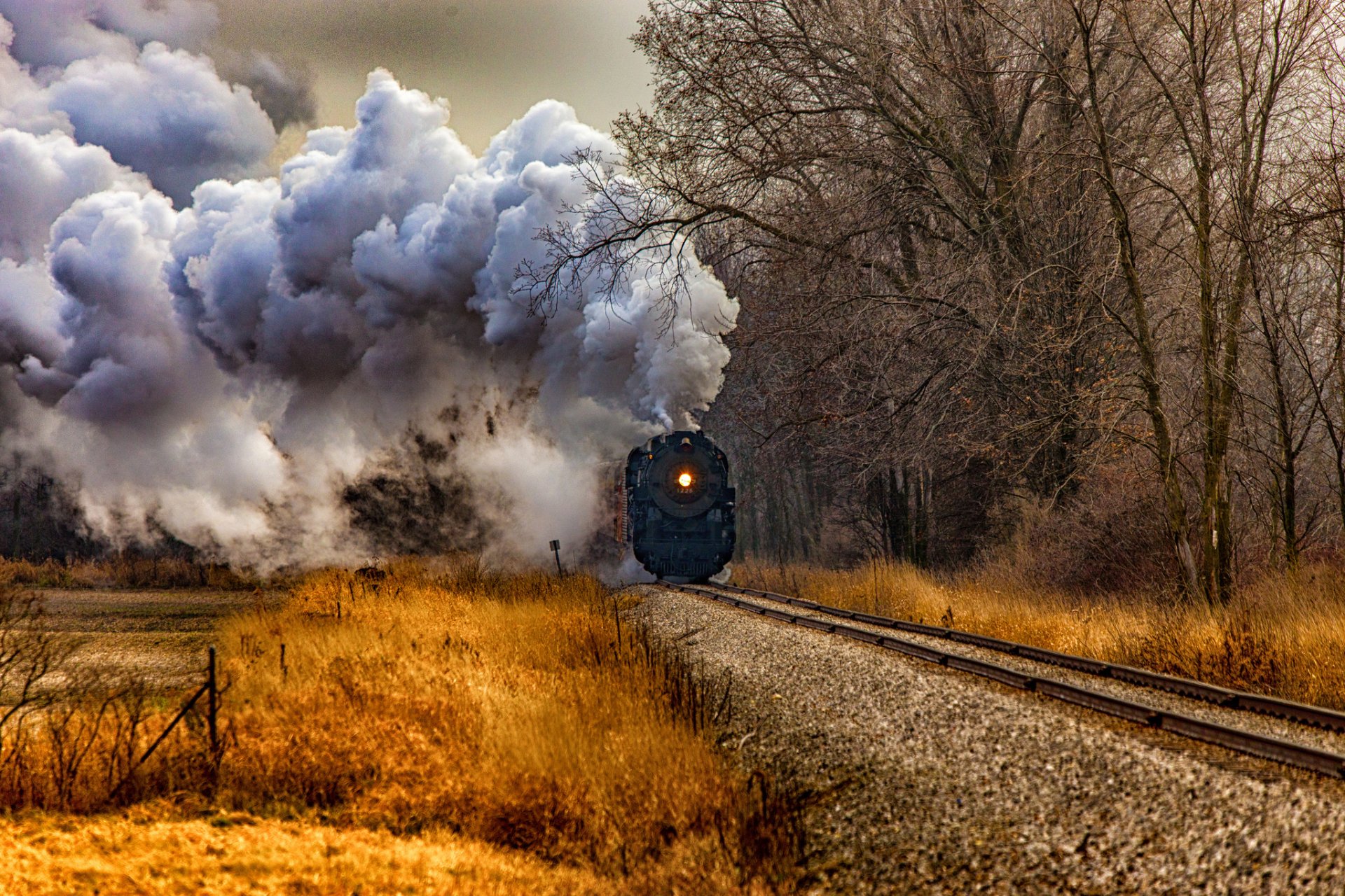 A classic locomotive billows smoke as it travels along a solitary track, surrounded by autumn foliage and trees, creating a dynamic scene for a HD desktop wallpaper.