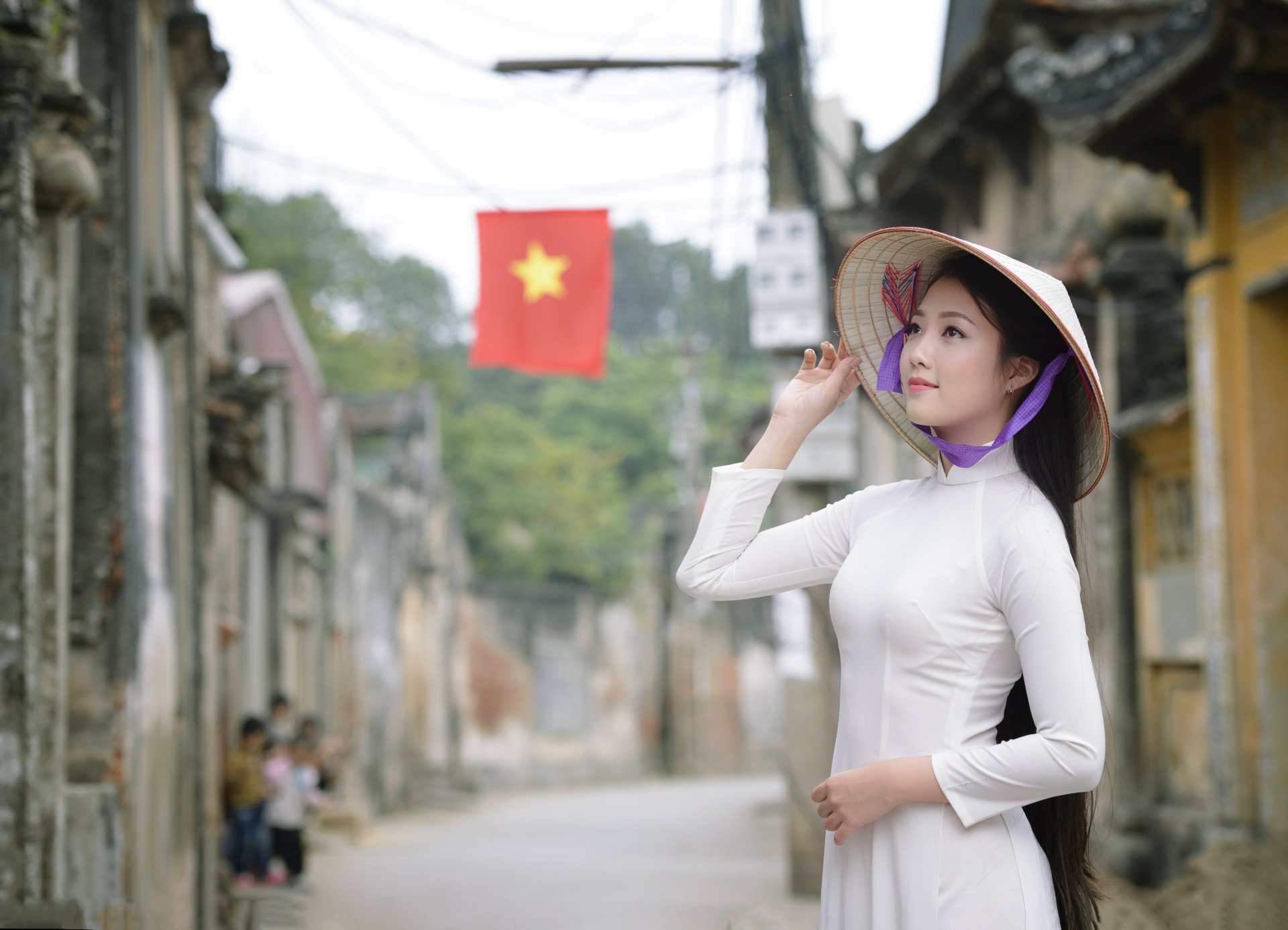 A woman in a traditional Ao Dai and conical hat poses gracefully on a charming street, with a Vietnamese flag in the background, capturing the essence of Vietnamese culture.