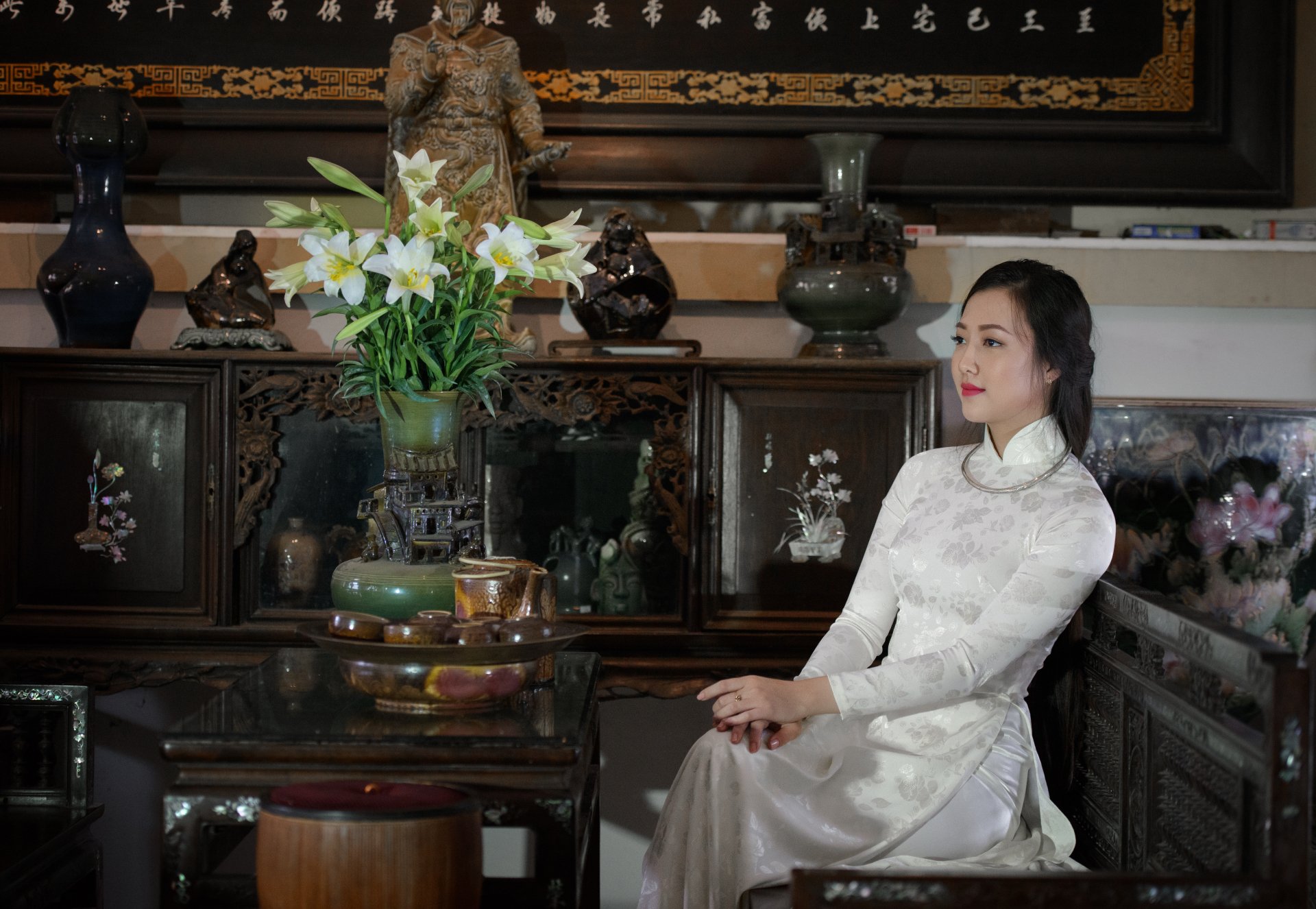 A Vietnamese woman in a traditional white Ao Dai sits gracefully beside a vase of lilies in an ornate, antique room, captured in 4K Ultra HD desktop wallpaper quality.