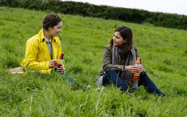 HD desktop wallpaper featuring Jenna Coleman and Emilia Clarke sitting on grass, each holding a drink, from the movie Me Before You.