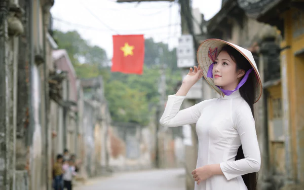 A woman in a traditional Ao Dai and conical hat poses gracefully on a charming street, with a Vietnamese flag in the background, capturing the essence of Vietnamese culture.