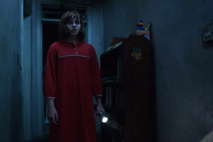 A young girl in a red nightgown holds a flashlight, standing apprehensively in a dimly lit hallway, from the movie *The Conjuring 2*. This eerie scene makes for an engaging HD desktop wallpaper.