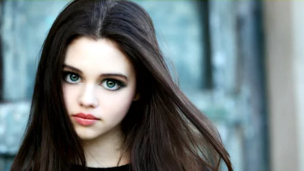 Close-up HD wallpaper of brunette child celebrity India Eisley with striking green eyes and flowing hair, set against a blurred outdoor background.