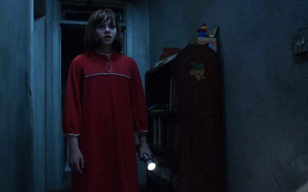A young girl in a red nightgown holds a flashlight, standing apprehensively in a dimly lit hallway, from the movie *The Conjuring 2*. This eerie scene makes for an engaging HD desktop wallpaper.