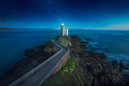 Evening view of a man-made lighthouse on the rocky coast of France, surrounded by the calm sea and ocean under a starry night sky.