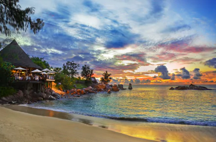 Tropical beach scene in Seychelles featuring a resort by the ocean, palm trees, and a stunning sunset horizon. Vibrant colors reflect on the water, creating a serene atmosphere.